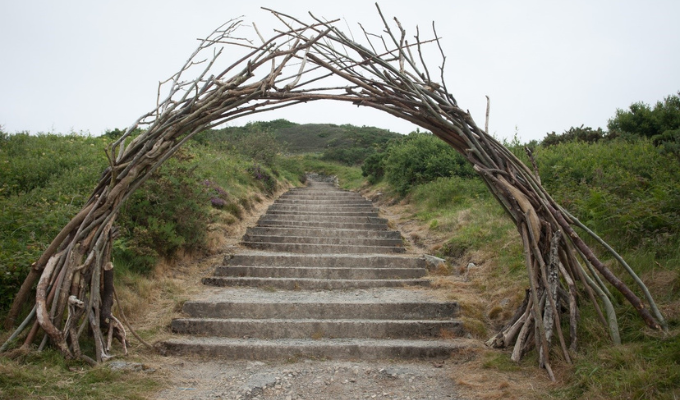 An archway made of tree sticks leads into a pathway of steps, with green fields on either side.